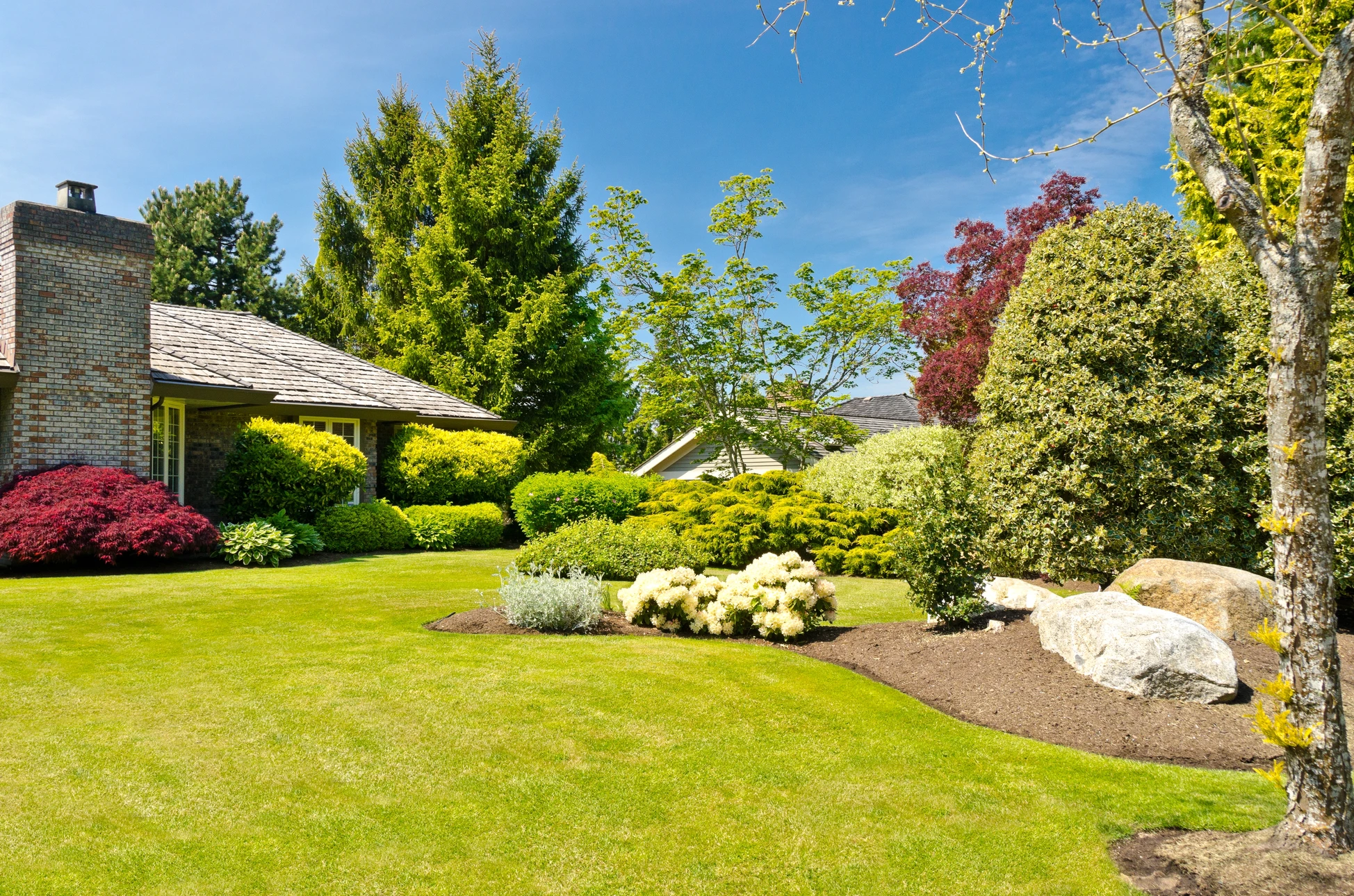 Photographie d'un jardin paysager contemporain aménagé avec expertise professionnelle, montrant une propriété résidentielle sophistiquée. Au premier plan, une terrasse en pierre naturelle calcaire ou composite de qualité, bien entretenue. À proximité, un parterre floral soigneusement composé associant végétation structurée et fleurs colorées. En arrière-plan, un espace verdoyant avec pelouse tondue impeccablement, massifs paysagers harmonieux, et possiblement une fontaine ou un bassin ornamental. L'image doit transmettre le professionnalisme, la qualité du travail et la transformation complète d'un espace extérieur par expertise paysagère. Lumière naturelle généreuse, ciel dégagé ou partiellement nuageux, composition permettant de bien voir l'ensemble du jardin aménagé en perspective.